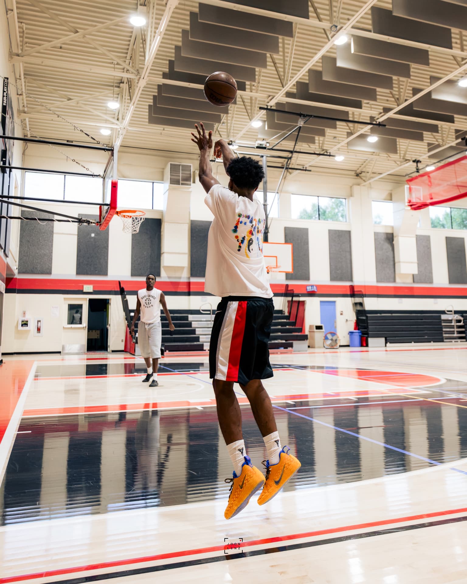 An athlete releasing a jump shot on an empty court while a coach watches from the baseline.